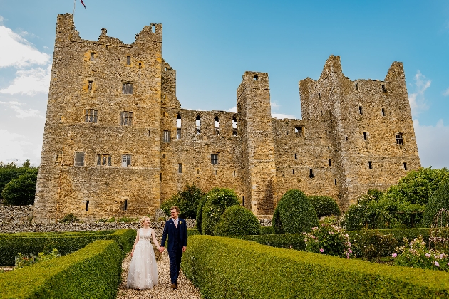 Bride and groom walk hand in hand in front of Bolton Castle